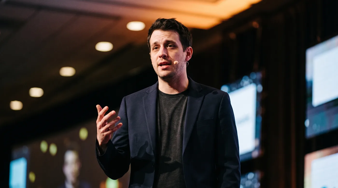 AI-edited photo of the same man speaking on a conference stage with blurred lights behind him.