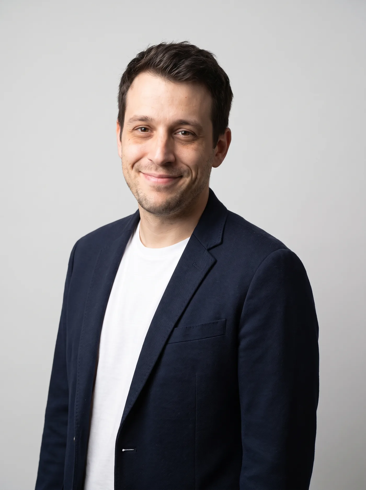 AI-edited corporate team-page portrait of the same man in a navy blazer against a plain grey studio background.