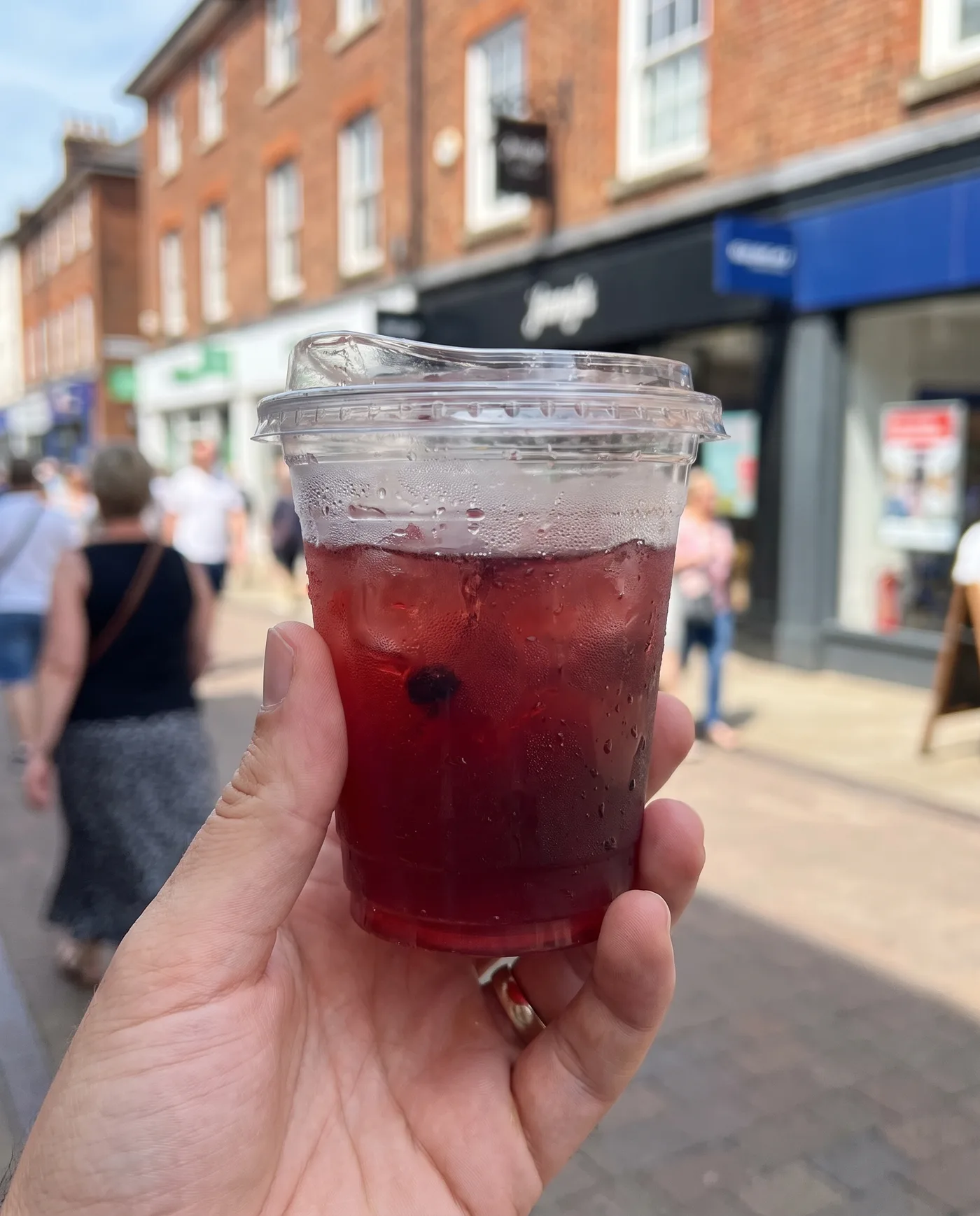 AI-generated casual phone-style photo of a hand holding an iced berry drink in a clear cup on a busy high street.