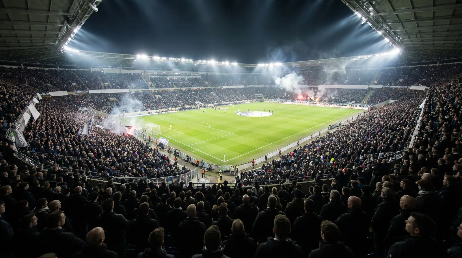 A packed football stadium on a matchday under floodlights, a sea of supporters filling the stands.